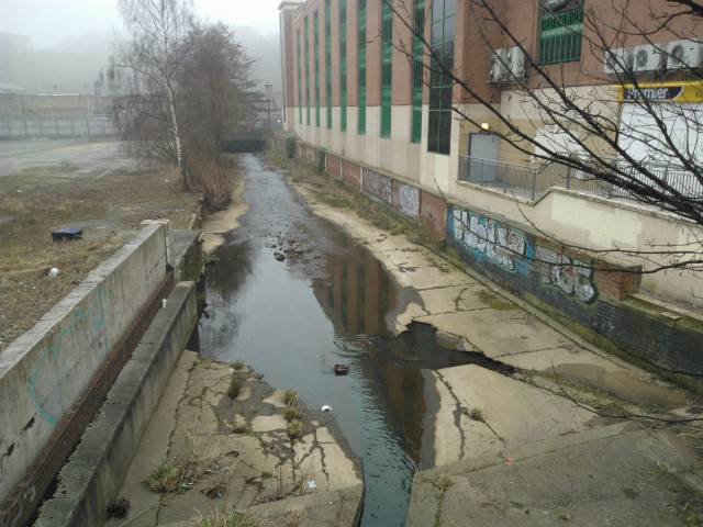 he last open section of the Porter Brook before it joins the River Sheaf in a culvert beneath Sheffield Station. It is degraded, concrete lined, and treated like a drain and rubbish tip. This is less than 100 metres from the fountains shown in the first picture. Image by author.